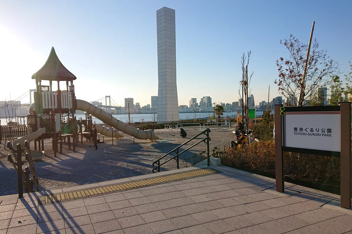 A children's playground with a slide and a tall pale tower at Toyosu Gururi Park in low evening sun, with Tokyo Bay and the Rainbow Bridge in the distance and a park sign on the right
