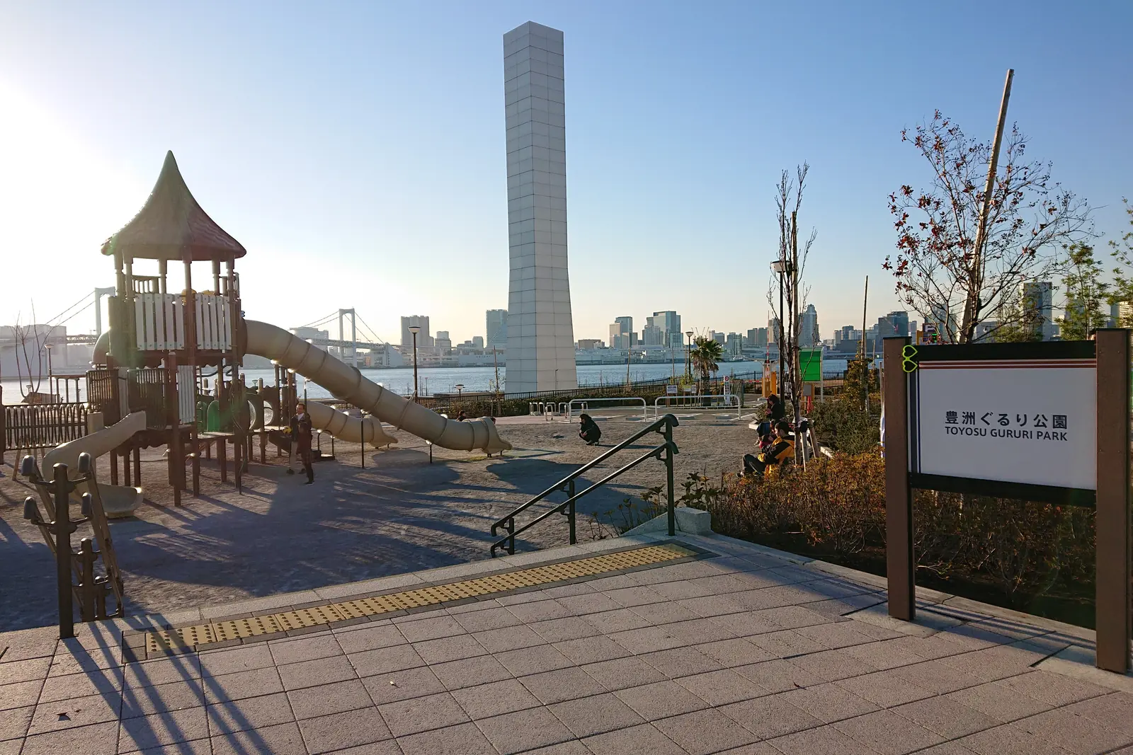 A playground with a slide and a tall pale tower at Toyosu Gururi Park in low sun, with Tokyo Bay and the Rainbow Bridge beyond; a general photograph of the Toyosu waterfront, not of the museum