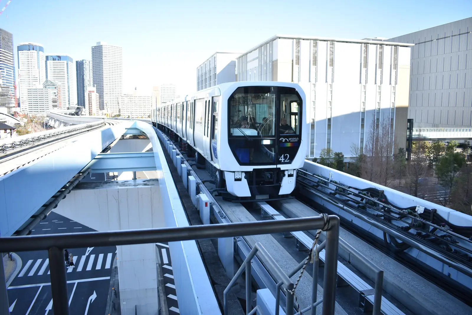 A white Yurikamome train on its elevated track above a street junction, with glass towers behind it at Shin-Toyosu Station